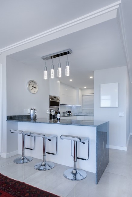 White and silver-themed kitchen, three silver counter stools, four turned-on hanging lights in the counter